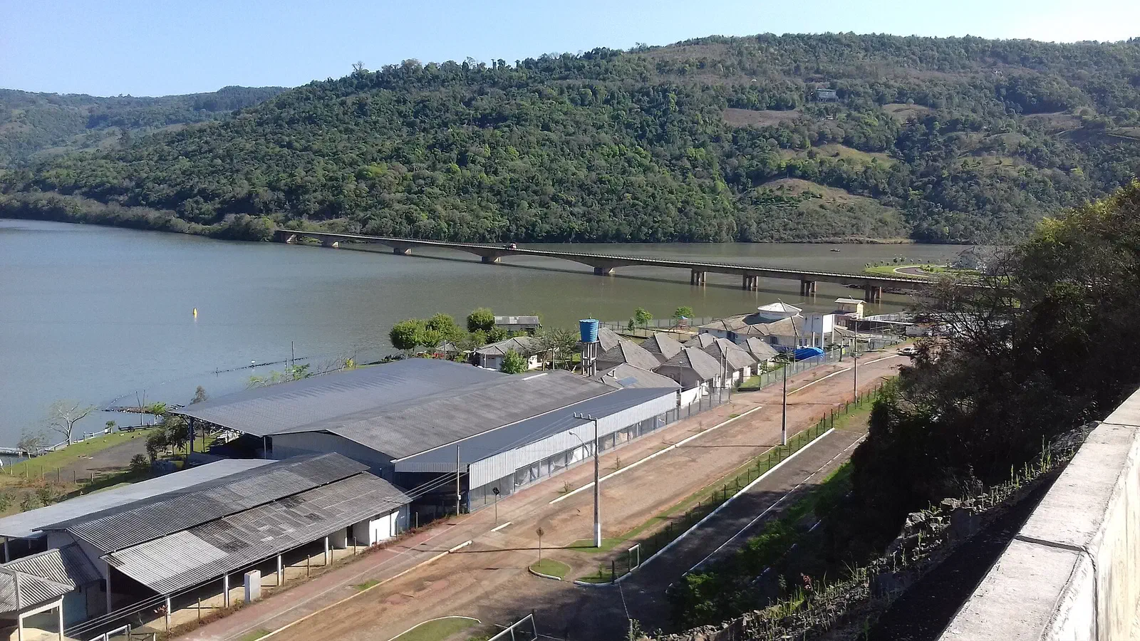 A ponte rodoviária sobre o rio Uruguai no Goio-Ên, herdeira da antiga travessia por balsa, vista de cima com as construções do distrito na margem catarinense e os morros de mata ao fundo