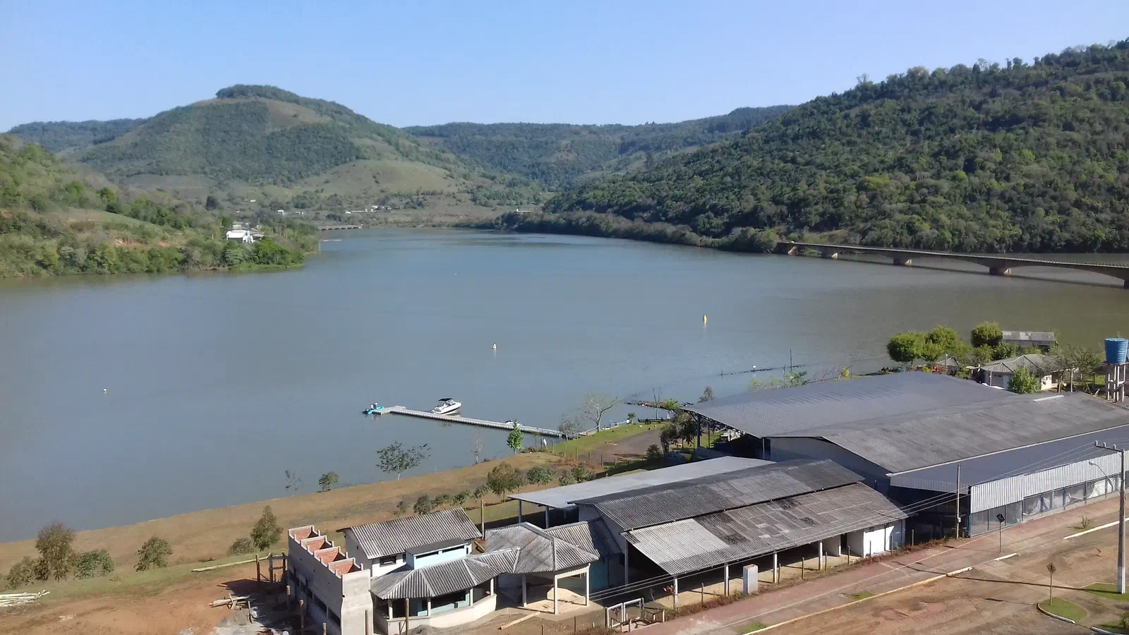 Vista do rio Uruguai no Goio-Ên, distrito de Chapecó/SC: o rio largo no vale entre morros cobertos de mata, com um trapiche na margem, a orla do distrito e a ponte rodoviária ao fundo à direita